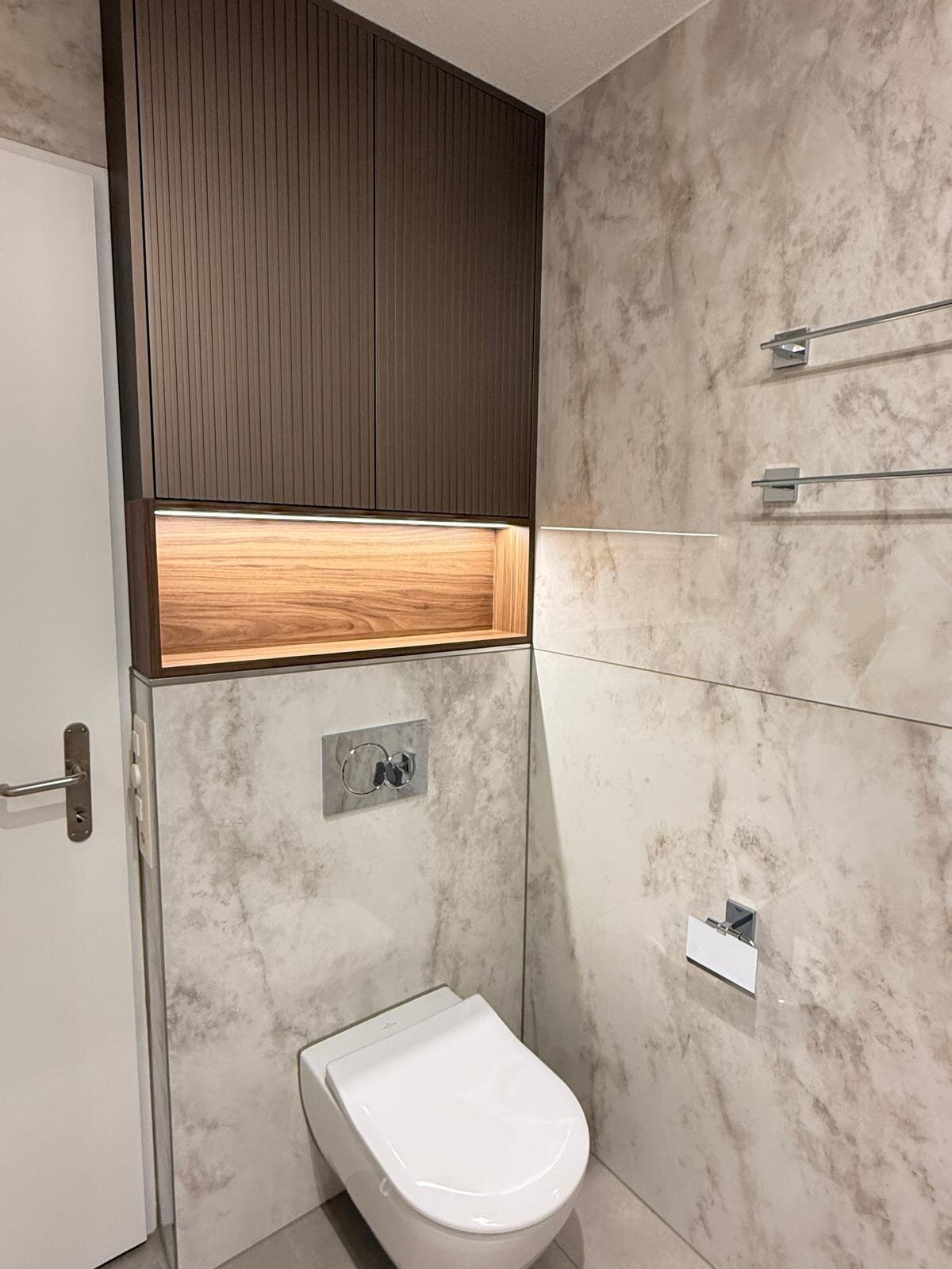 Wand-WC vor Naturstein in Marmor-Optik, darüber ein dunkles Holzpaneel mit beleuchteter Nische